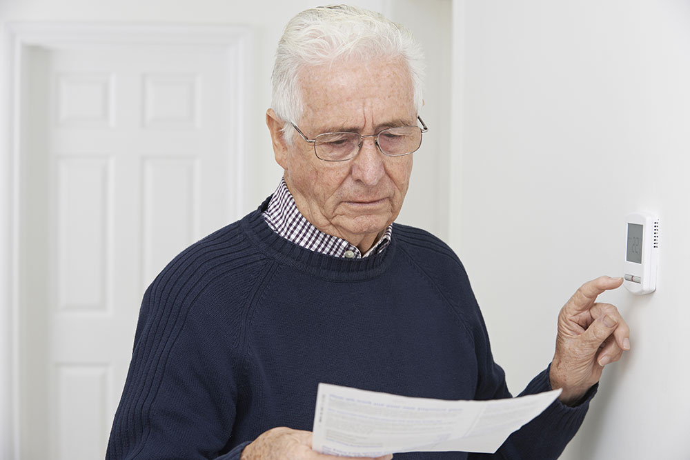 Elderly man looking at an electric bill while adjusting his thermostat