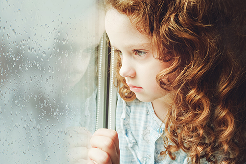 Sad redheaded little girl looking outside through a wet window